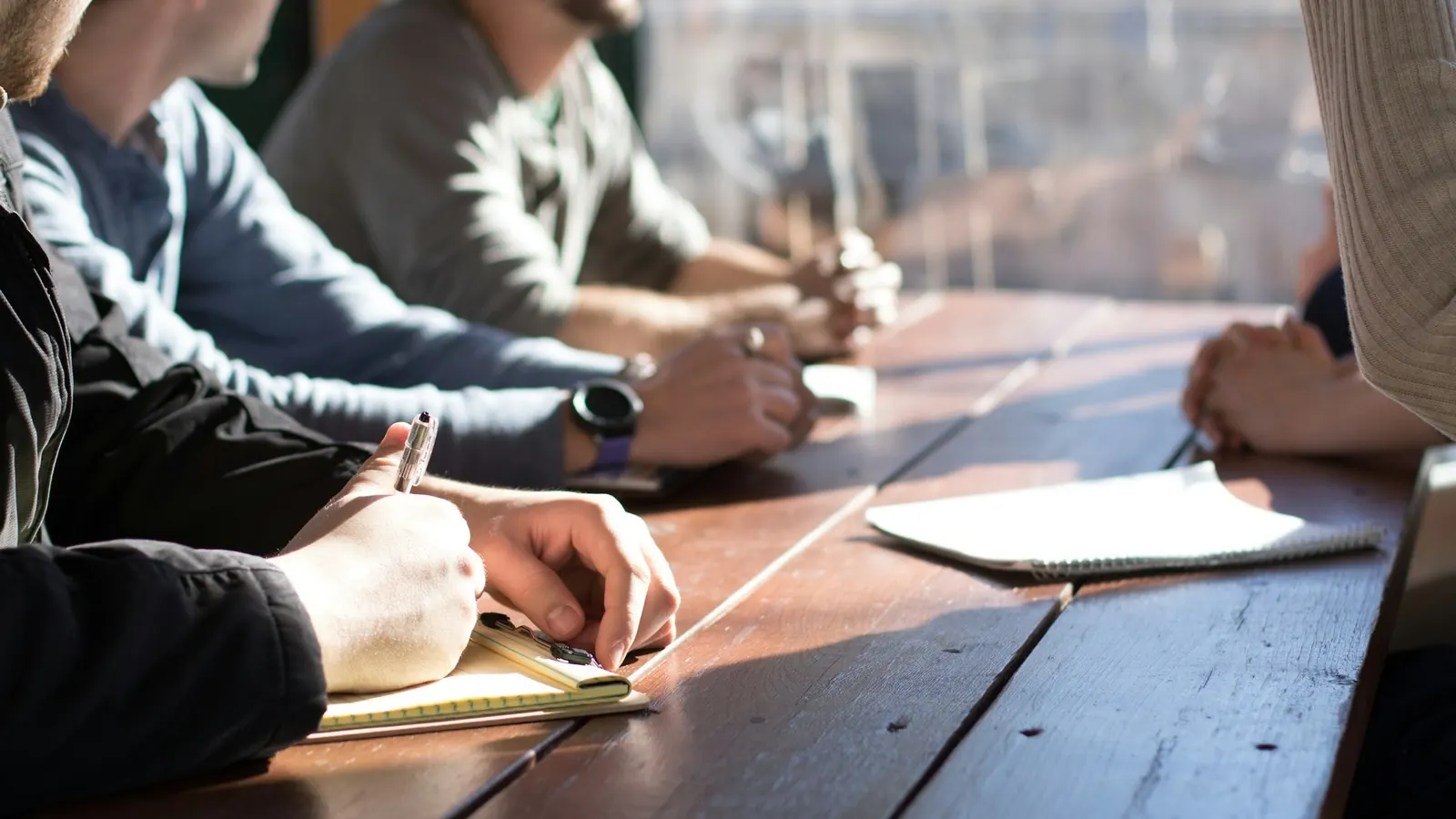 Réunion de travail autour d'une table en bois : une personne prend des notes sur un carnet, deux autres écoutent, mains posées sur la table